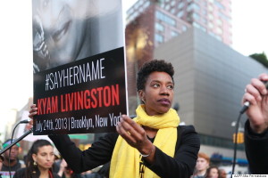 This image shows an African American woman protests on a city street. She stares off into the distance; her face reveals nothing. She is holding a sign that says hashtag say her name Kyam Livingston July 24th 2013 Brooklyn, New York. 