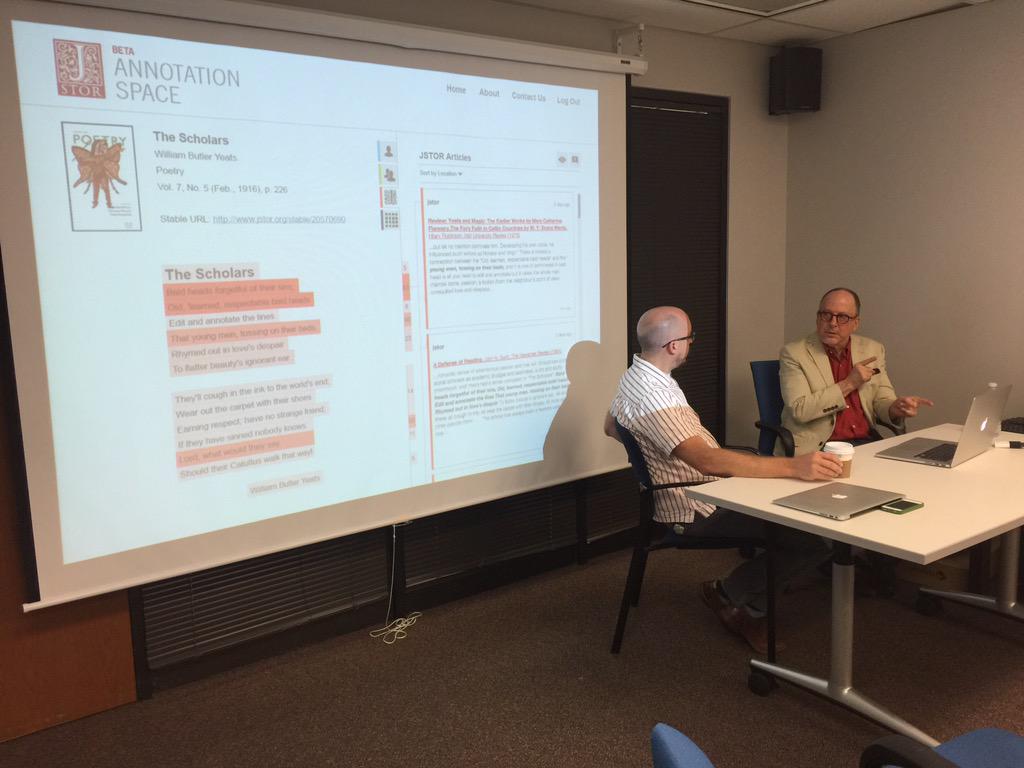 Two men in conversation over a laptop, sitting in front of a projector screen showing a poetry annotation screen.