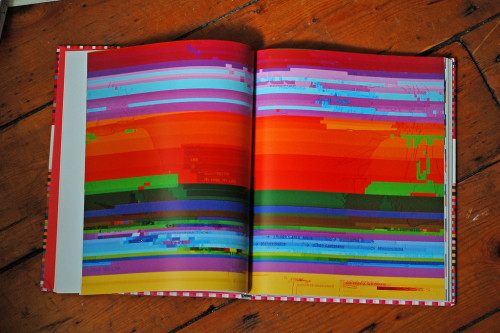 An open book in lying on a wooden floor. The pages show the bright color bands of computer screen displaying a visual glitch.