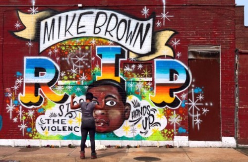 This image features a mural to honor shooting victim Michael Brown. It reads "MIKE BROWN. R. I. P. Stop the Violence. Hands Up." In front of the mural, a young black woman stands with her back to the camera. Her hands are raised.