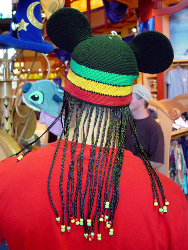 From-behind view of a white man in a Disney store. He is wearing a hat with Mickey Mouse ears, stripes in the African colors green, yellow and white, and fake black-haired braids dangling down his neck.