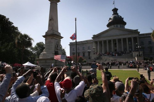 Image shows a crowd watching the Confederate Flag be lowered outside a building with columns.