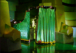 Approximately four second gif from the movie The Wizard of Oz. The image starts with a little dog biting the bottom of a green curtain. The dog then pulls the curtain back to reveal a man wearing a suit and standing in an enclosed space. His back is toward the camera and he's unaware the dog has pulled back the curtain. The man is working an unknown technical device. He pulls on levers while lights on the machine blink.