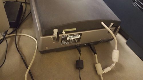 This photo is of the back of a scanner. It shows the power and USB cords plugged into the scanner. Both ports are toward the middle of the scanner and about an inch apart. The power cord is on the left, and the USB is to the right.