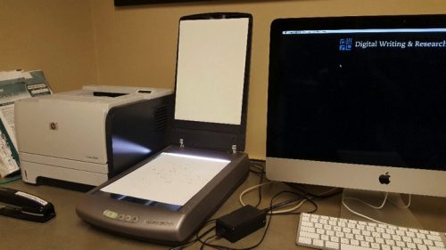 Similar to the photo from the third slide. There's a HP printer (on the left), an Epson scanner (middle), and a Mac computer monitor and keyboard. In this photo, however, the scanner's lid is lifted up and there is a white sheet of paper face-down on the scanner glass.