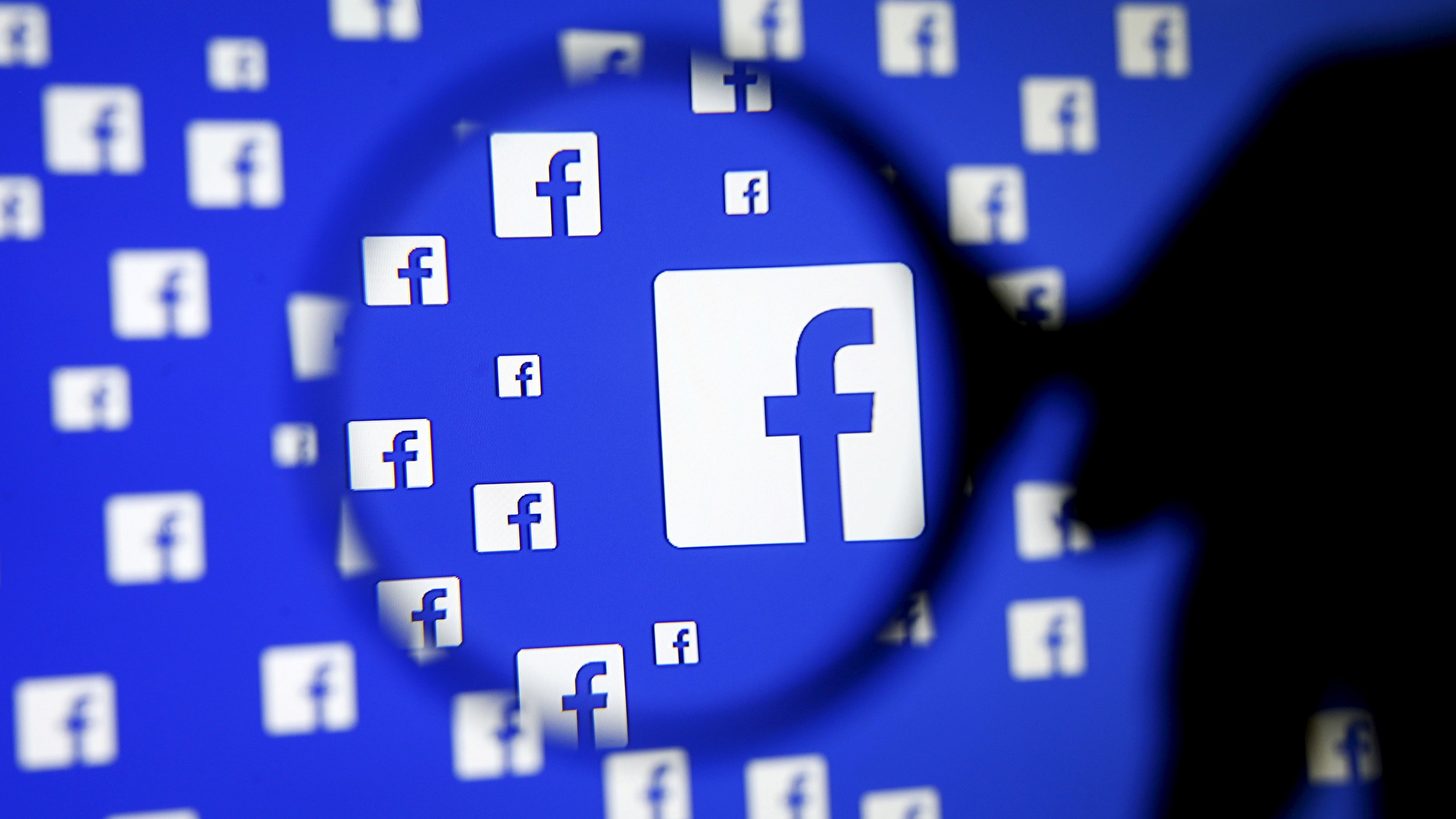 Soft-focus sillhouette of a magnifying-glass over a blue screen displaying many copies of the Facebook logo