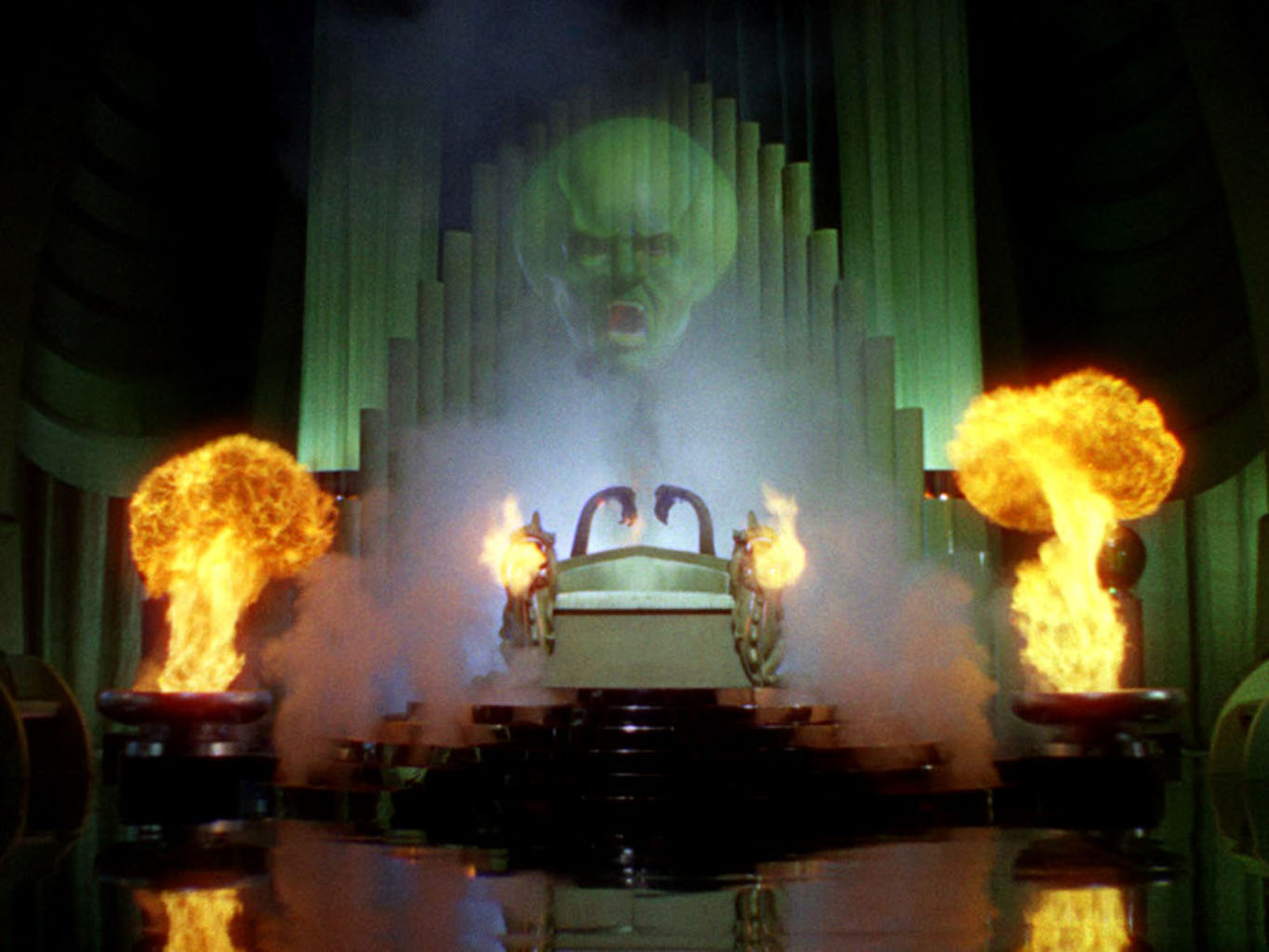 The image shows the intimidating, green, and translucent head of a man hovering in the air above some sort of altar in what could be a throne room. The man is shouting, and he has an enlarged cranium. Below the floating head is dense smoke. Partially encased by the smoke is a metalic scultupre with fire coming out of its sides toward the camera. At both the left and the right edges of the altar, fire erupts out of short but thick columns. The floor is black, but shiny and reflective. The back of the altar (behind the semi-transparent head) are tall narrow columns that resemble a massive church organ. 