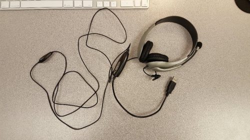 This picture is of a microphone/headphone headset lying on top of a computer desk. Attached to the headset is a long black cord, at the end of which is a USB plug. 