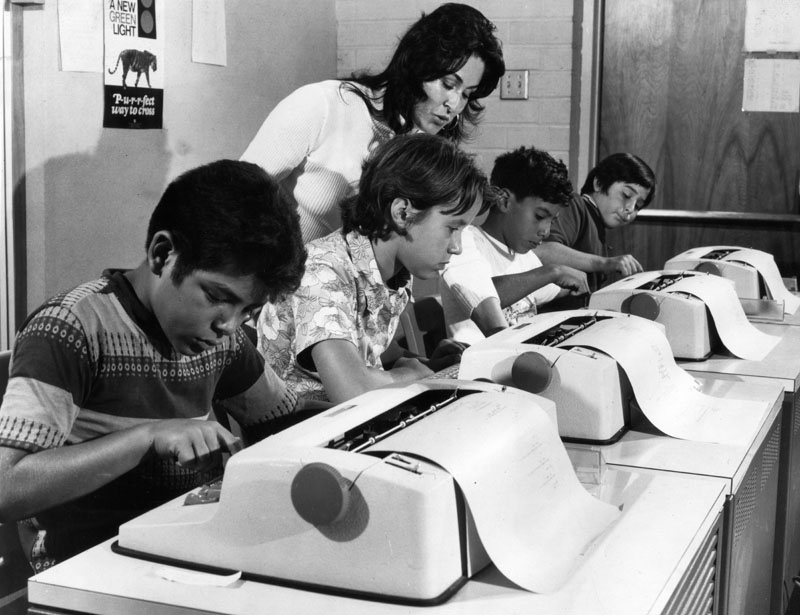 Black and white photo with a teacher standing behind four children and typrewriter like computer terminals.