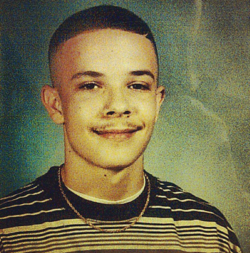 This image reproduces the high school yearbook photo of Shaun King. He is smiling, with a light mustache and an edge up haircut. He is wearing a black and white T-shirt and a gold necklace.