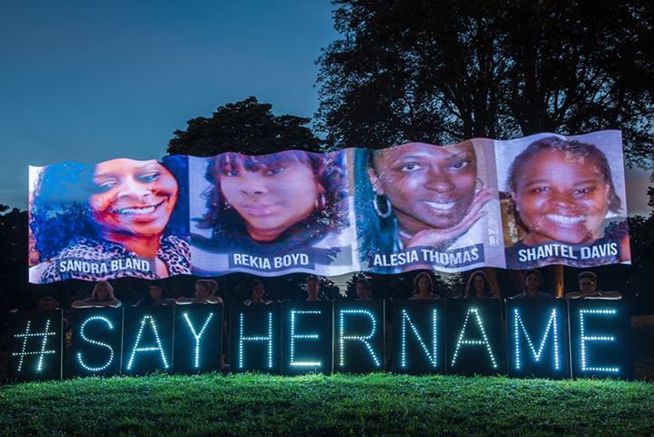 illuminated text "#sayhername" in a park with photographs of four black women: Sandra Bland, Rekia Boyd, Alesia Thomas, Shantel Davis