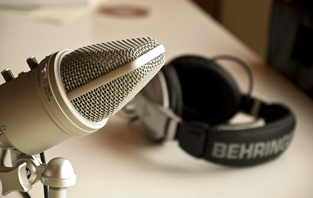 A close-up photograph of a studio microphone. The microphone extends into the left half of the frame. In the background on the right, and out of focus, is a set of Behringer headphones lying on a white table.