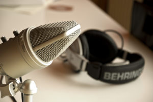 A close-up photograph of a studio microphone. The microphone extends into the left half of the frame. In the background on the right, and out of focus, is a set of Behringer headphones lying on a white table.