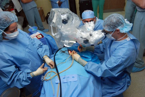 Three surgeons in blue lab coats crowded around a patient and performing open heart surgery. The patient is entirely covered by a blanket or coat in the same color as the surgeons'.