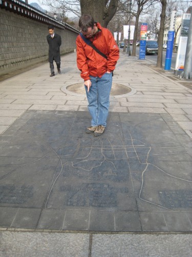 A man is standing on top of a map that is set into the pavement of a sidewalk. He is bent over, looking down on the map. The index finger of his right hand points somewhere on the map, but it is not clear where.