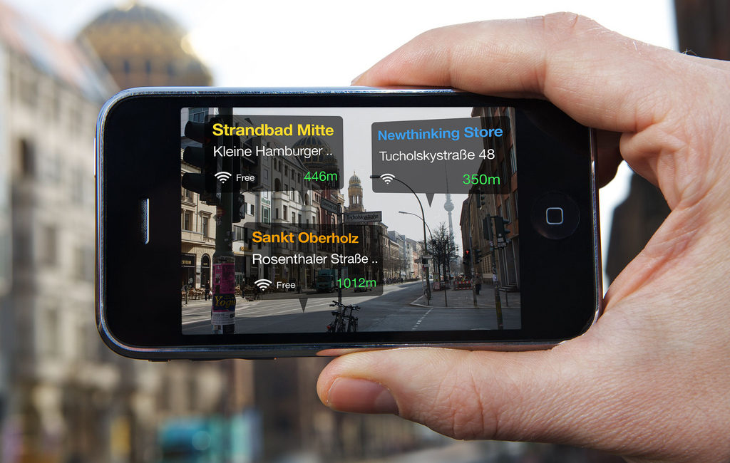 A hand is holding up a smartphone that shows an AR-app in use. The screen looks like the camera mode is on, but there are three translucent boxes overlaid on the streetview, which show the names of three locations on a German street that have free wifi, as well as their wifi's signal strength and the walking distance to each location.