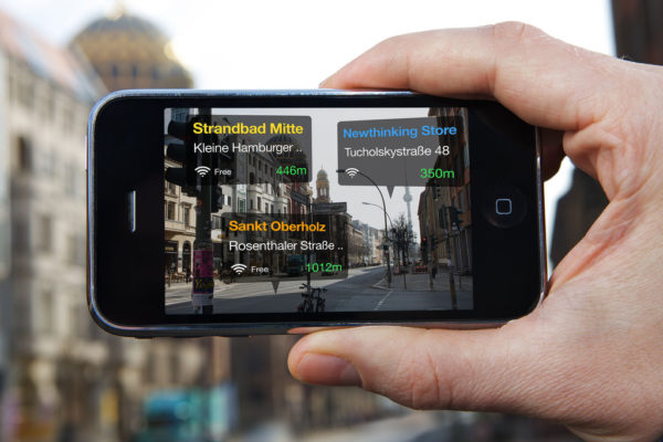 A hand is holding up a smartphone that shows an AR-app in use. The screen looks like the camera mode is on, but there are three translucent boxes overlaid on the streetview, which show the names of three locations on a German street that have free wifi, as well as their wifi's signal strength and the walking distance to each location.