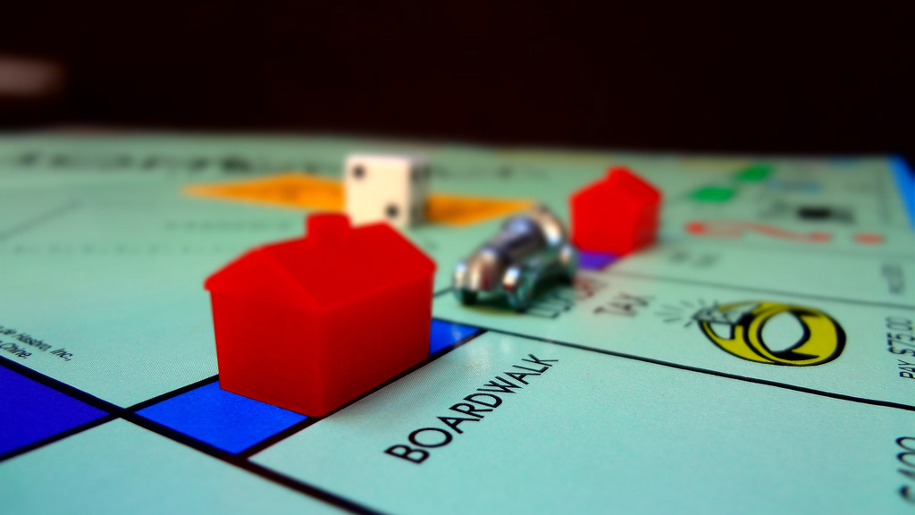 The corner of a Monopoly gameboard showing the space "Boardwalk," which is a high value property in the game. The space also has a hotel on it, which indicates that it is a lucrative property. Behind the plastic, red hotel is a small metal car gamepiece.