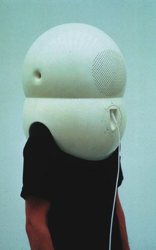 A picture of a man wearing a giant white device over his head. The device is plugged into something out of view, and the device looks extremely inconvenient and cumbersome. The image conveys a sense of devices taking their users hostage, or humans being inconvenienced by that which they designed for the sake of convenience