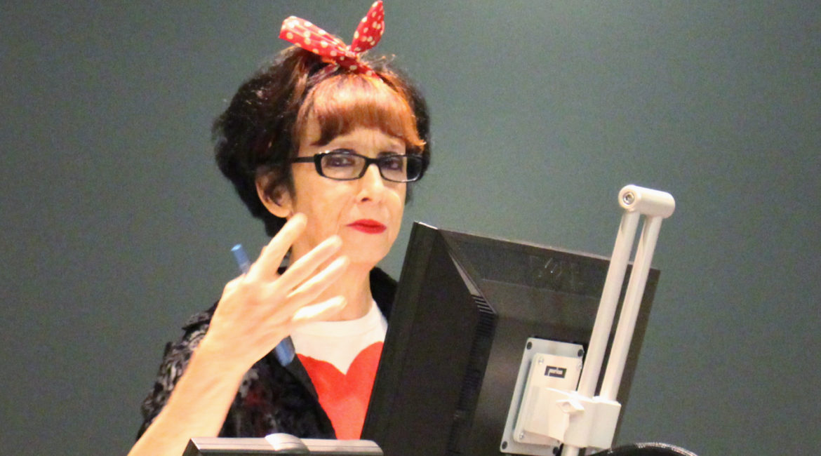 This is a picture of Avital Ronell in a speaking gesture with her right arm, spreading her four fingers and holding a blue pen with her thumb. She is standing in fron of two computer screens. Behind here is a dark wall. She is looking into the camera and is wearing black rectangular glasses. She has red lipstick and a red ribbon in her her. Her hair is mainly black, and she wears a black jacket with a white-red blouse.