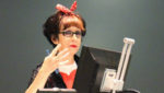 This is a picture of Avital Ronell in a speaking gesture with her right arm, spreading her four fingers and holding a blue pen with her thumb. She is standing in fron of two computer screens. Behind here is a dark wall. She is looking into the camera and is wearing black rectangular glasses. She has red lipstick and a red ribbon in her her. Her hair is mainly black, and she wears a black jacket with a white-red blouse.