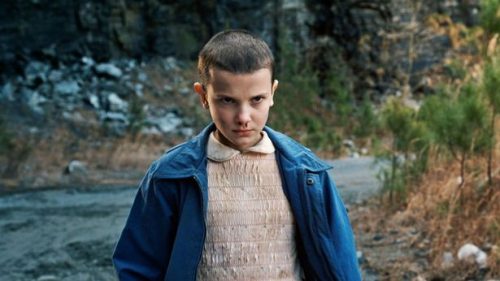 An young girl, maybe twelve years old, stands on a dirt road with a rockface in the background. The photo shows her from the waste up, and she's wearing a dirty pink blouse and a blue coat. Her hair is short, her nose is bleeding, and her look is intense.