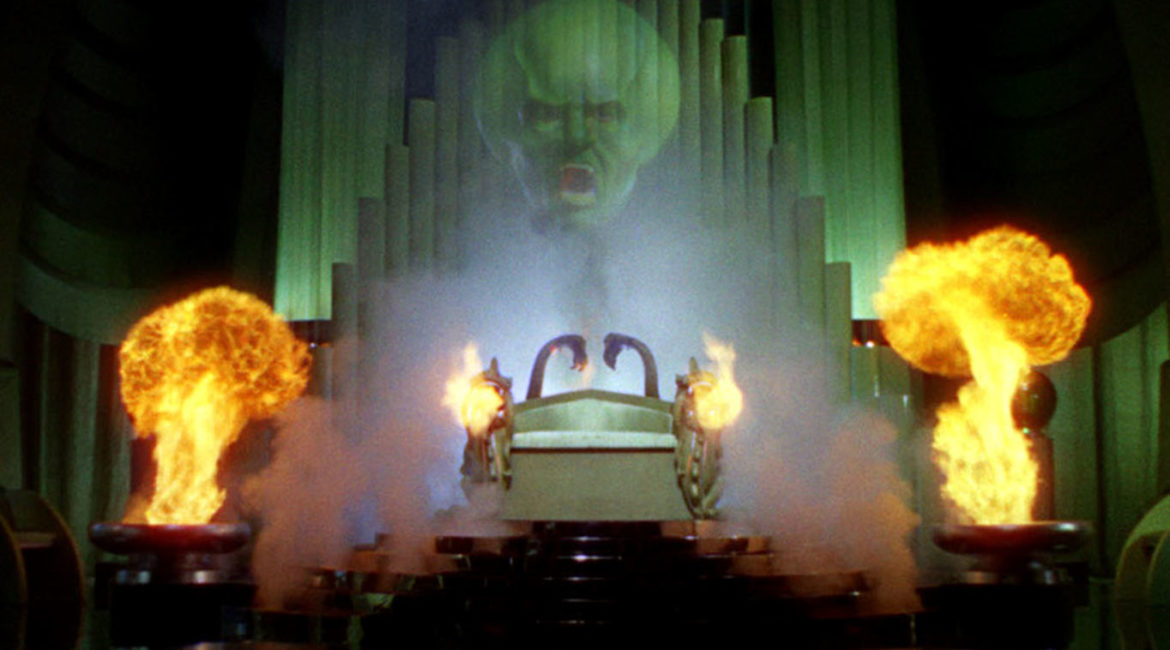 The image shows the intimidating, green, and translucent head of a man hovering in the air above some sort of altar in what could be a throne room. The man is shouting, and he has an enlarged cranium. Below the floating head is dense smoke. Partially encased by the smoke is a metalic scultupre with fire coming out of its sides toward the camera. At both the left and the right edges of the altar, fire erupts out of short but thick columns. The floor is black, but shiny and reflective. The back of the altar (behind the semi-transparent head) are tall narrow columns that resemble a massive church organ.