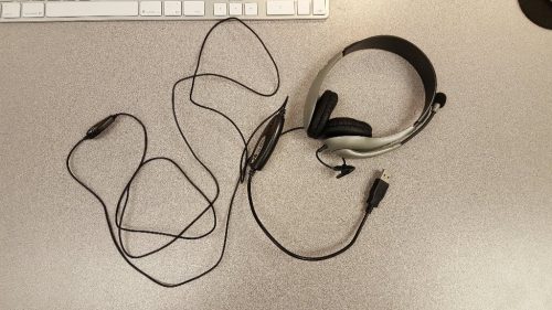 This picture is of a microphone/headphone headset lying on top of a computer desk. Attached to the headset is a long black cord, at the end of which is a USB plug.