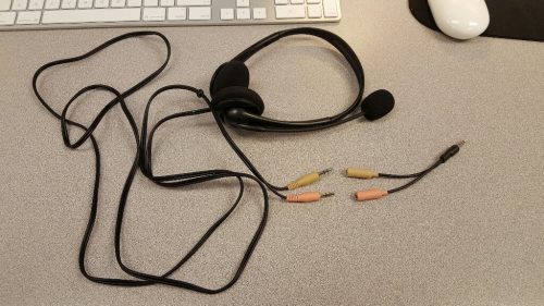 This picture is of a black headphone/microphone headset lying on a computer table. Attached to the headset is a long black cord that leads to two different plugs (one yellow and the other pink). Near those is a Y-shaped adapter, with two jacks that match the two plugs. At the other end of the adapter is a single metalic plug. 