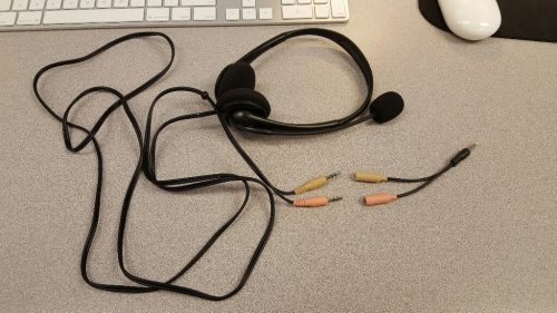 This picture is of a black headphone/microphone headset lying on a computer table. Attached to the headset is a long black cord that leads to two different plugs (one yellow and the other pink). Near those is a Y-shaped adapter, with two jacks that match the two plugs. At the other end of the adapter is a single metalic plug.