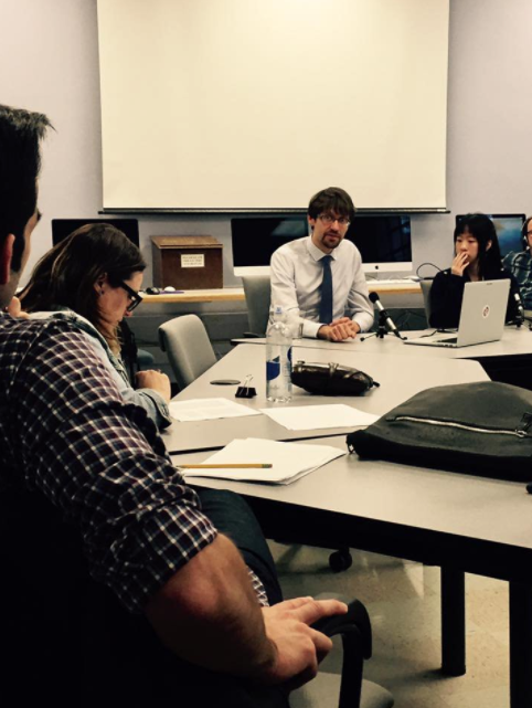 Picture of people at a curved table looking towards a man in a blue shirt and tie speaking, it is a picture from when Professor Patrick Jagoda Hosted a Q&A with DWRL Staff