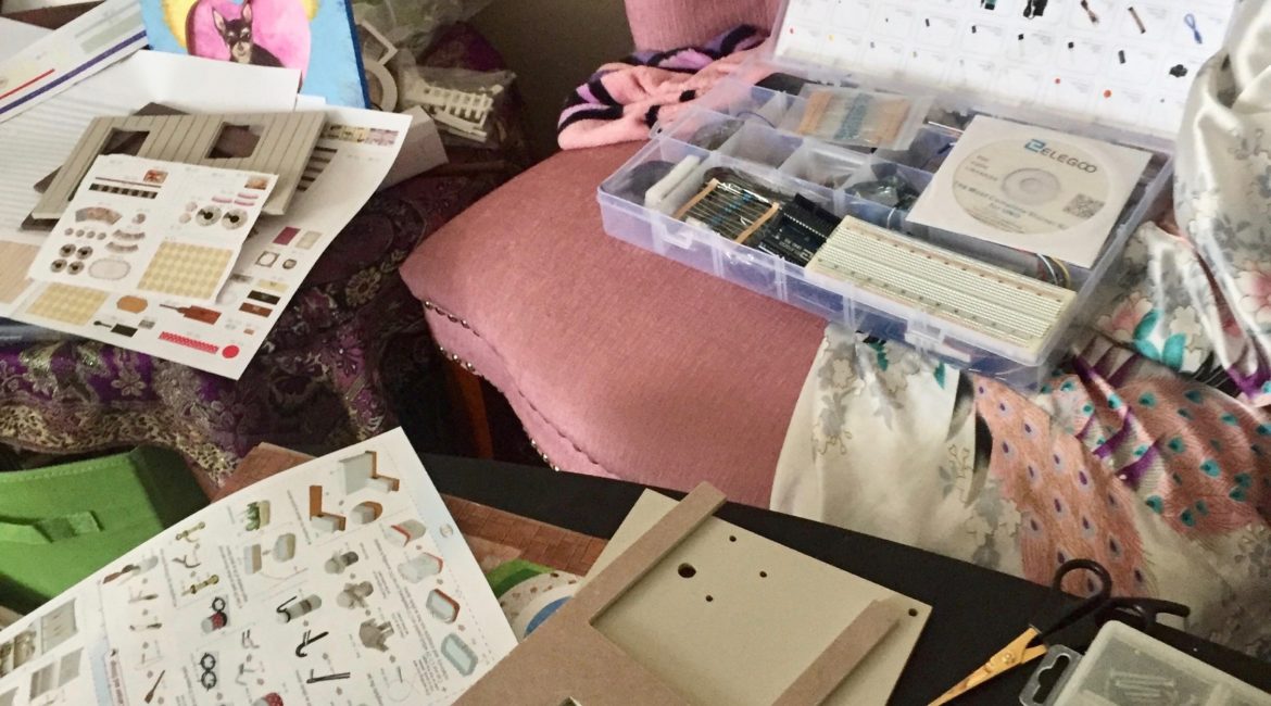 An Arduino computing kit sits atop a plush, pale pink chair in the background of the image. In the foreground, various supplies and instructions for a model home rest next to a pink glue gun on a black table.