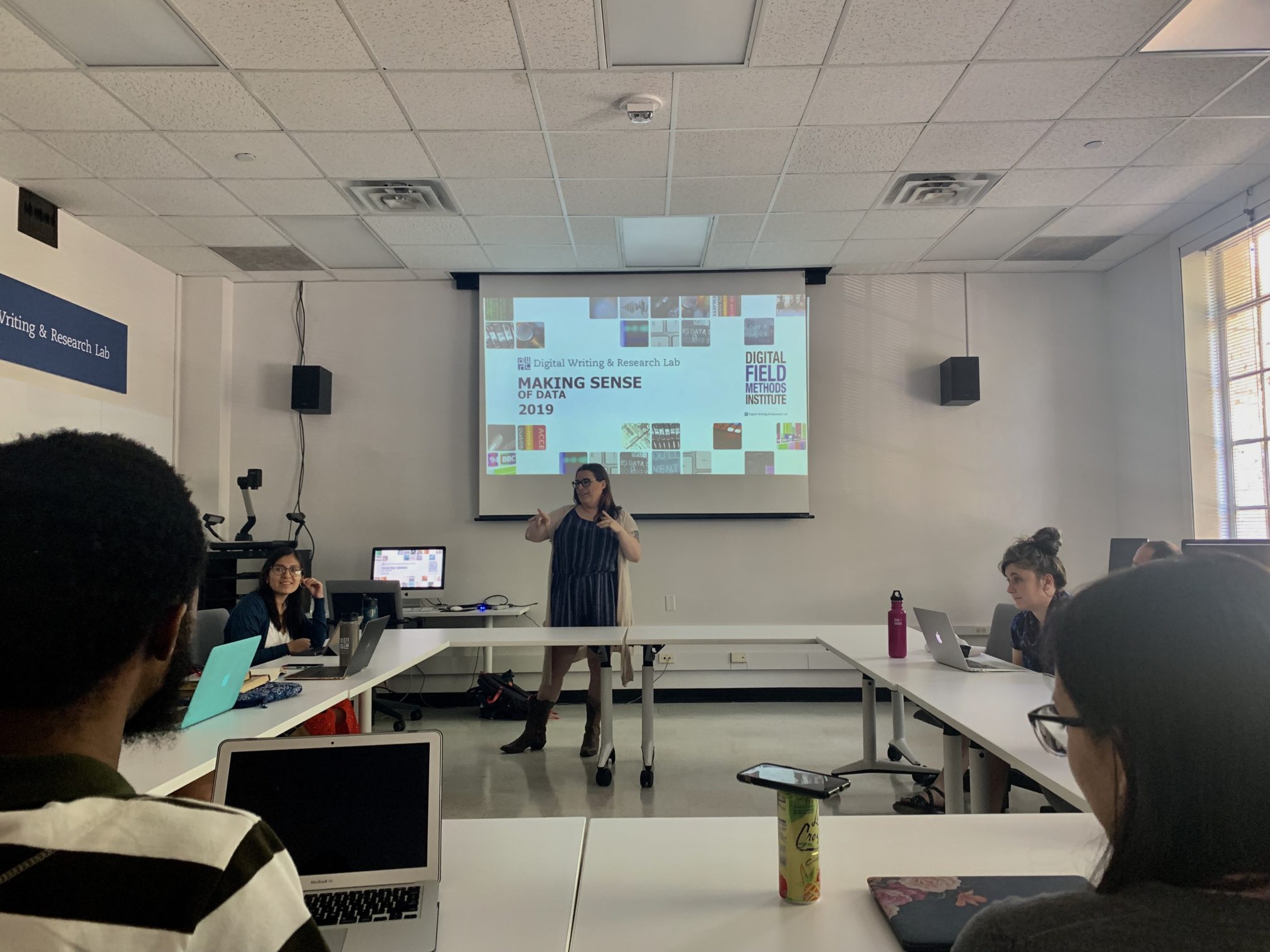 A woman stands in front of a screen presenting to a group of people. The screen reads "Making Sense of Data"